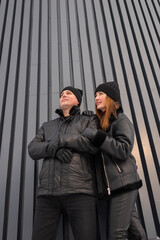 Fashionforward Pair In Dark Clothing Poses Near Architectural Background, Two Individuals Donning Black Jackets Stand Confidently Against Contemporary Striped Wall Showcasing Urban Style