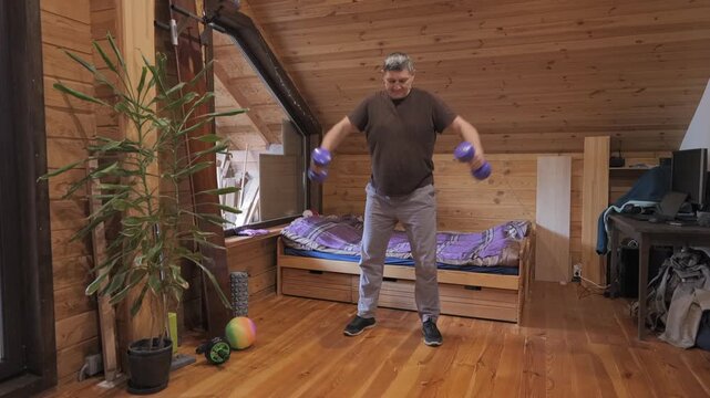 Middle aged man performing dumbbell lateral raises inside wooden attic room, indoor strength exercise session using hand weights during personal home fitness practice