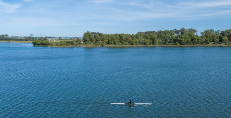 Aerial view of a lone rower gliding across the expansive, serene blue lake, framed by lush green trees, Ciudad de la Costa, Departamento de Canelones, Uruguay.