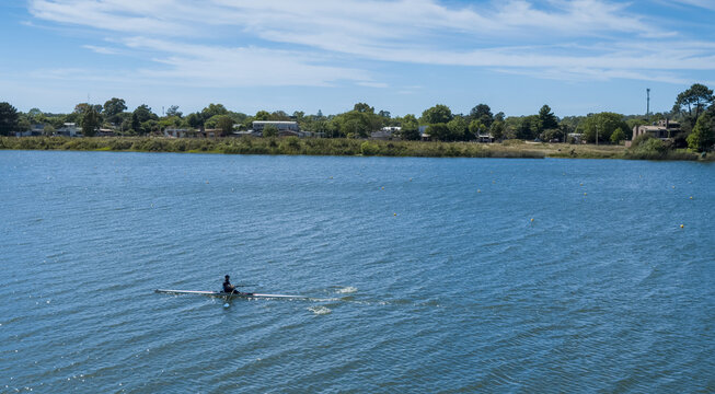 Aerial view of a lone rower gliding across the tranquil lake waters reflecting the clear sky, near lush green shores, Ciudad de la Costa, Departamento de Canelones, Uruguay.