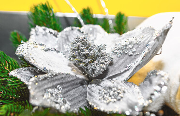 Glittering silver artificial flower with sequined petals rests among green pine branches, evoking Christmas festive charm. Bright and soft white tones in background enhance its decorative appeal