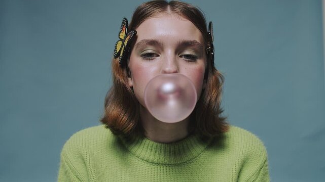 Young woman with butterfly hair clips blowing large pink bubble gum bubble against blue background. Playful trendy mood suitable for youth lifestyle and fashion content.