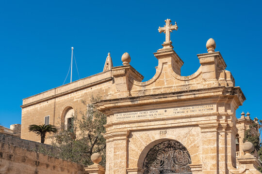 View of an ornate stone gate adorned with a cross, leading to Parish Church of Mellieha under a clear sky, casting shadows, Mellieha, Malta.