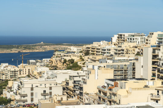View of sun-drenched buildings cascade down towards the azure sea, a distant island beckons under the clear sky, Mellieha, Malta.