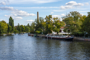 View of calm waters reflecting the sky and surrounding trees, with a boat docked near a brick building with a tall chimney, Koepenick, Berlin, Germany.
