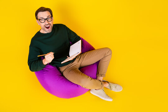 Young man in green sweater sits on a purple beanbag writes in a notebook smiling against a bright yellow background for lifestyle fashion and casual advertising