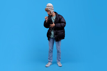Full body of young Asian man in winter clothing coughing on blue background