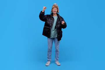 Happy Asian young man celebrating success in winter clothing against blue background