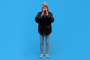 Young Asian man in winter clothing shouting with hands cupped around mouth against blue background