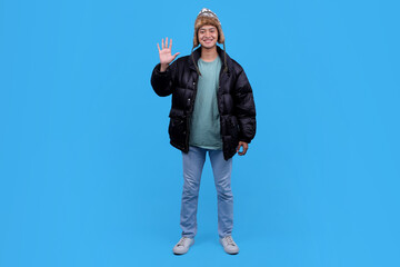 Full body young Asian man waving hand in winter hat and black puffer jacket on blue background