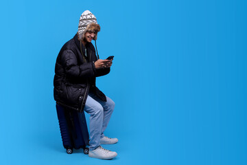 Young man in winter clothes sitting on luggage with phone in hand