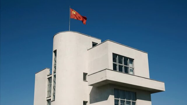 Red flag with hammer and sickle above a white modernist building. Historical architecture in former USSR territory. Symbol of communism.