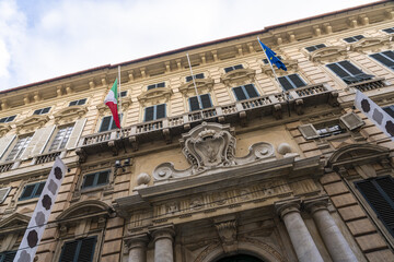 Genoa, Italy - 25 December 2022: View of a grand building facade adorned with flags, intricate carvings, and closed shutters under a cloudy sky.