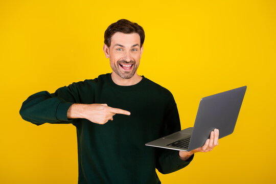 Young man holds a laptop smiling and pointing at the screen against a bright yellow background - Powered by Adobe