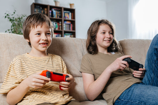Siblings playing video games with console controllers on sofa at home
