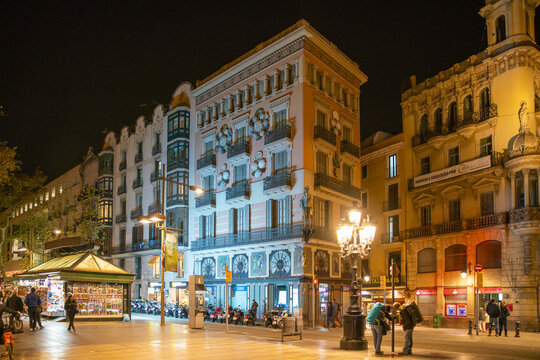 Barcelona, Spain - 07 November 2016: View of illuminated buildings casting warm light onto the bustling Placa de Catalunya, where people gather beneath vintage street lamps in the heart of the city.