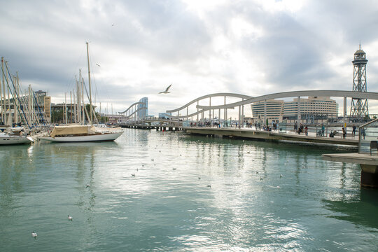 Barcelona, Spain - 07 November 2016: View of the tranquil harbor waters reflecting the sky, boats, and the iconic Rambla de Mar bridge against a cloudy skyline.