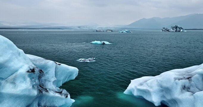 Massive blue icebergs frame a narrow channel of cold seawater in Vatnaj&ouml;kull lagoon, with smaller bergs drifting toward distant mountains under a grey winter sky.