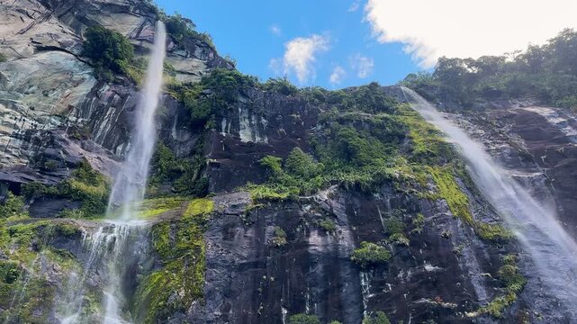 Looking up at towering twin waterfalls in Milford Sound, Fiordland National Park, New Zealand. Misty cliffs, cascading water, and dramatic fjord landscapes create a breathtaking scene.