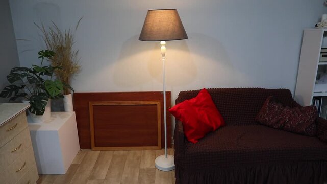 Smart floor lamp near a small sofa. Interior decoration with a plant, chest of drawers and empty picture frame. Home comfort concept.