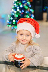 A cute girl with a mug at the table, a Christmas tree and a fireplace create a cozy festive atmosphere.