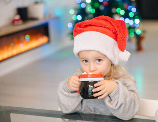 A cute girl with a mug at the table, a Christmas tree and a fireplace create a cozy festive atmosphere.