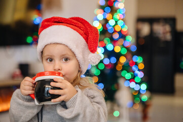 A cute girl with a mug at the table, a Christmas tree and a fireplace create a cozy festive atmosphere.