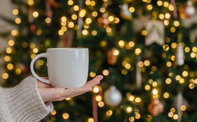 Hand in cozy sweater holding white mug with glowing Christmas lights behind.