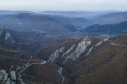 Aerial view of winding roads curl through rugged peaks and valleys, a tapestry of autumn hues fading into the distant blue mountains, Pietrapertosa, Basilicata, Italy.