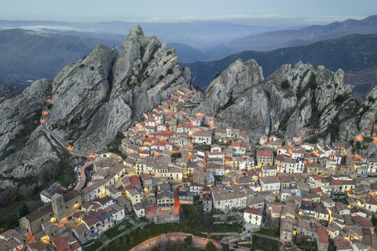 Aerial view of a village nestled against jagged, grey rock formations, lights twinkling against the deepening twilight, a harmonious blend of nature and architecture, Pietrapertosa, Basilicata, Italy.