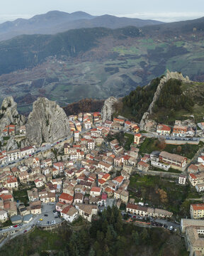 Aerial view of a village nestled amongst sharp, jagged rock formations, terracotta rooftops contrasting with the grey stone and distant green mountains, Pietrapertosa, Basilicata, Italy.