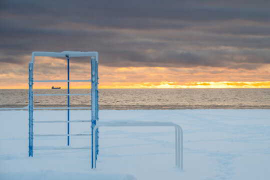 View of icy playground equipment stands starkly against the serene Baltic Sea, illuminated by a golden sunset under a moody sky, Parnu, Estonia.