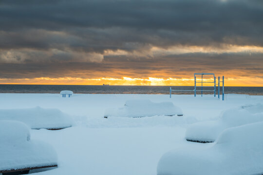 View of snow-covered benches and playground equipment under a dramatic sky with a glimpse of sunlight over the Baltic sea, Parnu, Estonia.