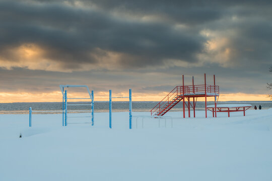View of red metal stairs and blue metal bars on a snow-covered beach under a cloudy, golden-tinged sky, Baltic Sea, Parnu, Estonia.