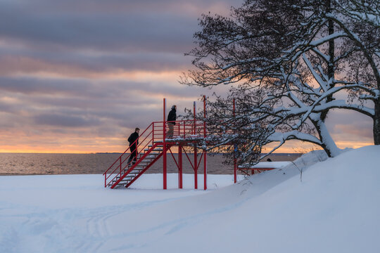 View of a vibrant red staircase contrasting against the stark white snow and the pale winter sky, with people silhouetted against the horizon, Parnu, Estonia.