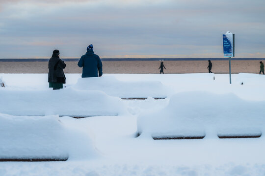 View of snow-covered benches and distant figures line the shore under a soft, overcast sky, creating a serene winter scene in Parnu, Estonia.