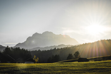 View of the morning sun casting a golden glow across the grassy field, silhouetting the dark forest and majestic mountain range in Krun, Bavaria, Germany.