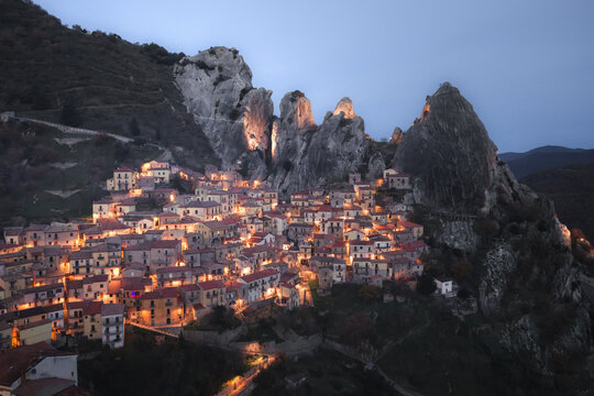 Aerial view of the lights of a village nestled between towering, craggy peaks glow warmly against the deepening twilight, Castelmezzano, Basilicata, Italy.