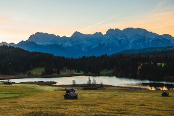 View of alpine lake mirroring the snow-dusted mountain range under a pastel sky, fringed by a dark forest and dotted with rustic huts, Krun, Germany.
