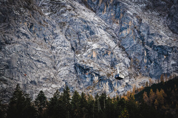 View of a cable car ascends dramatically against the rugged, textured face of a towering mountain, juxtaposed with the dark green forest below, Garmisch-Partenkirchen, Bavaria, Germany.