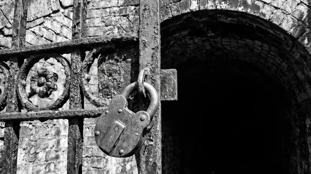 Ornate iron gate locked on ancient stone archway. Victorian era architecture detail with floral design and padlock. Historic building entrance secured in black and white vintage style.