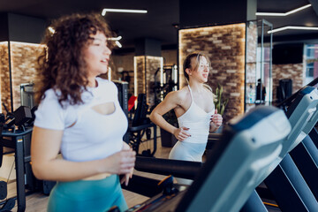 Side view of two beautiful young women in stylish sportswear running side by side on treadmills 