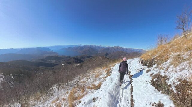 Monte Lema, Varese, Italy: Hiker walks on a narrow snow trail halfway up the mountainside in front of a breathtaking panorama of mountains and lake