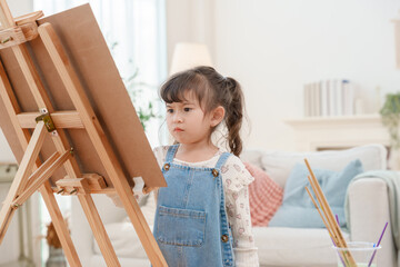Serious little girl concentrating on painting at wooden easel in living room creative hobby and child development concept