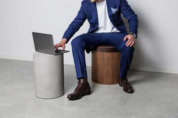 A man in a blue suit and white shirt leans forward to use a laptop on a concrete pedestal, seated...