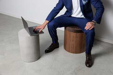 Man in blue suit seated on a modern stool, reaching to type on a laptop placed on a concrete pedestal. Minimalist setting with ample copy space