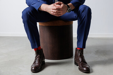 A man in a sharp blue suit sits on a dark wooden stool with hands clasped, looking to the side in a...
