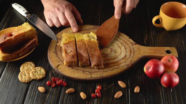 A person sorts pieces of cake on a wooden cutting board. Fresh apples, nuts, and cookies lie nearby. A cup of drink is visible on the table. The atmosphere is very cozy.