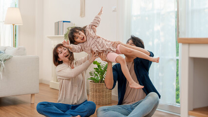 A delighted Asian mother and father lifting their cheerful young daughter high in the air, simulating flight during playtime in their brightly lit living room, embodying family happiness.