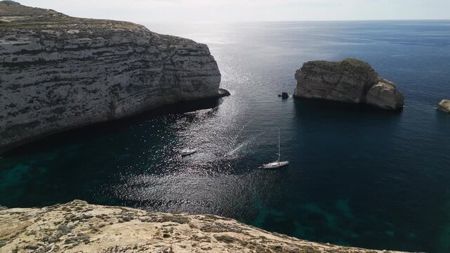 rone aerial view of sailboat anchored in protected bay at Dwejra, Gozo, Malta, cinematic 4K 60FPS footage showcasing dramatic limestone cliffs and Mediterranean seascape